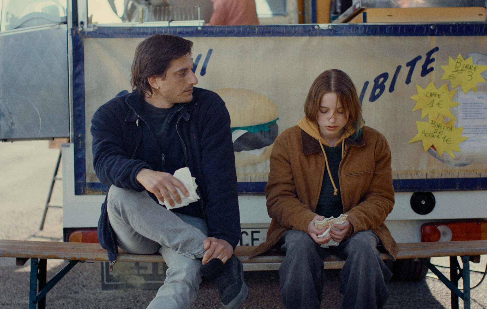 Ein erwachesener Mann und eine Teenagerin sitzen auf einer Bank. Er schaut sie an, sie schaut auf den Boden. Sie haben beide ein Sandwich in der Hand.