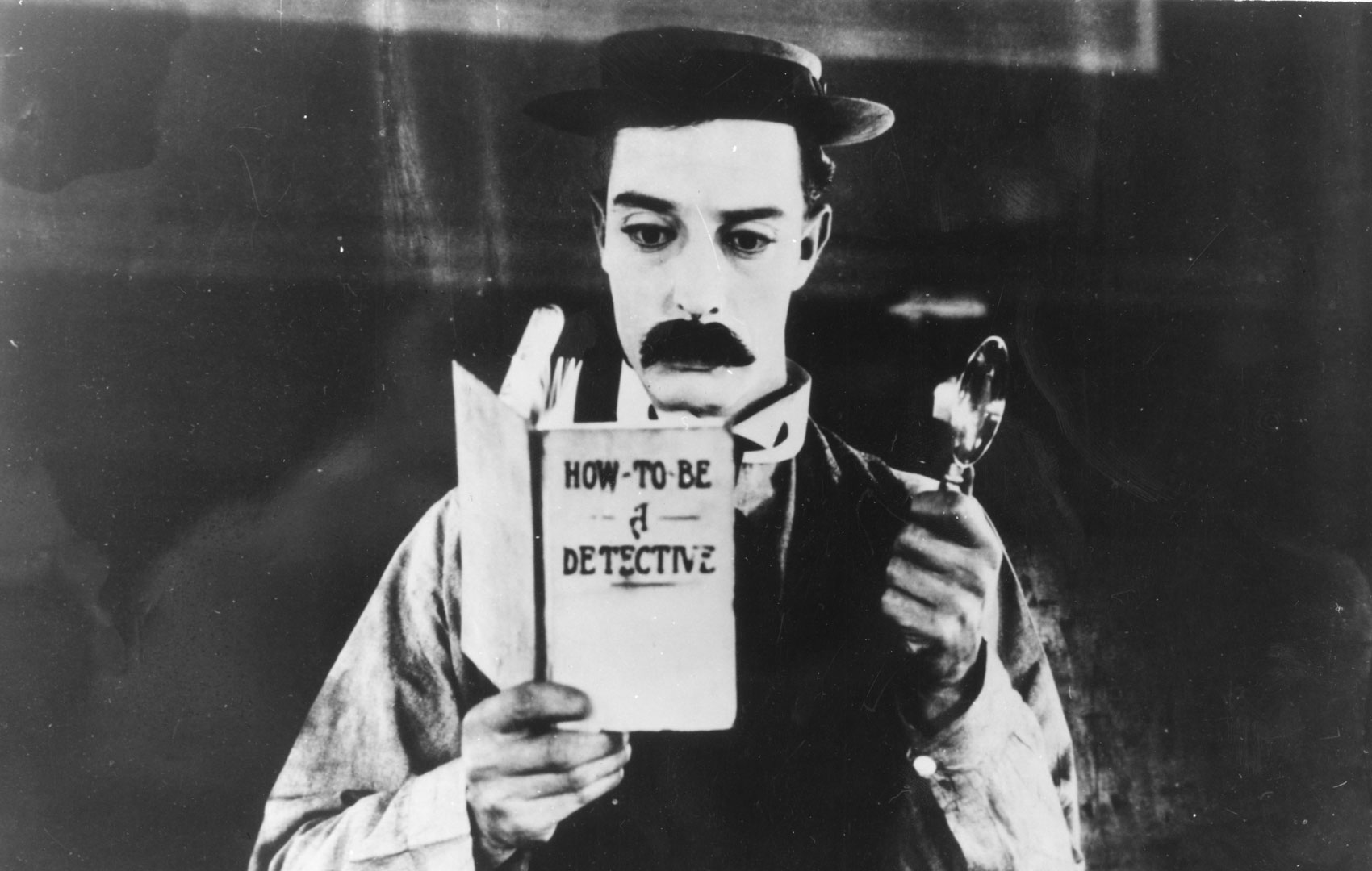 Schwarz-weiß Bild: Ein Mann mit Schnauzer und flaschem Hut hält in der einen Hand eine Lupe nach oben und mit der anderen Hand ein Buch. Dieses heißt: HOW TO BE A DETECTIVE