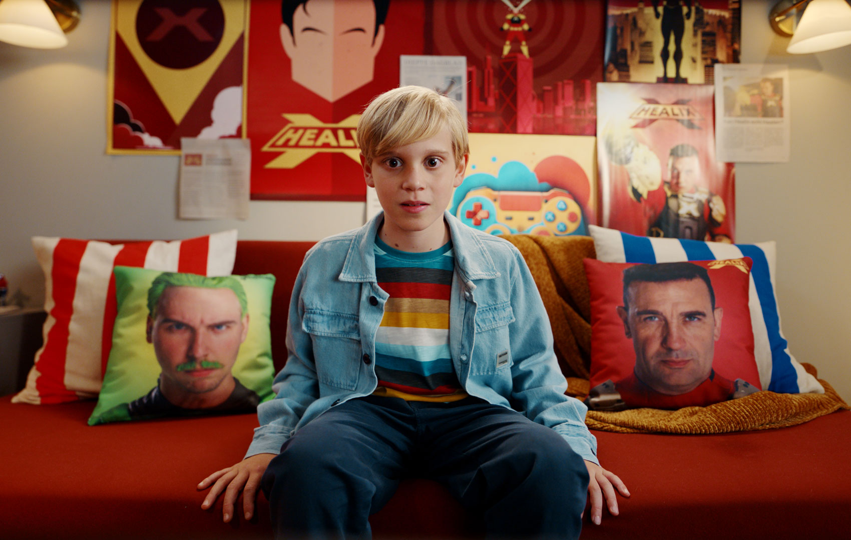 Ein Junge sitzt mit entgeistertem, frontalen Blick in die Kamera auf einem roten Sofa. Rechts und links neben ihm liegen Kissen, auf die Bilder von Männerköpfen gedruckt sind sind.