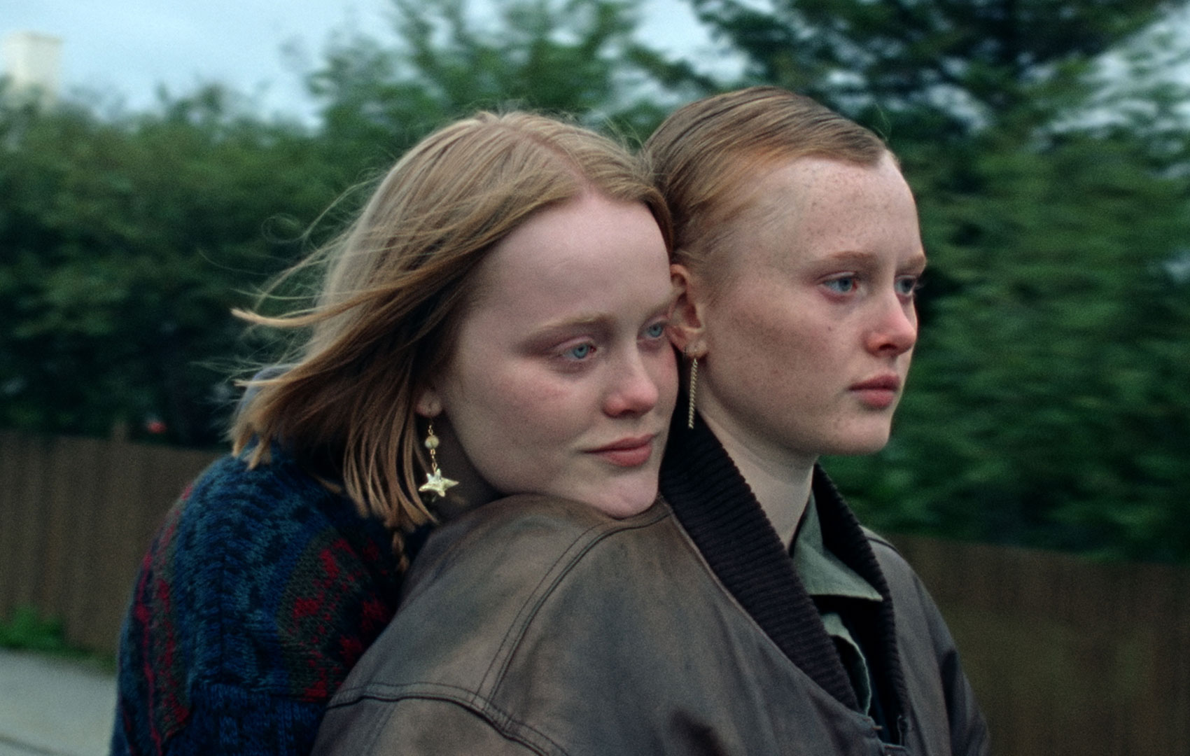 Eine junge Frau hat ihren Kopf auf der Schulter einer anderen Frau abgelegt. Sie schauen beide in die gleiche Richtung nach rechts aus dem Bild heraus. Sie sind draußen. Im Hintergrund sieht man einen Holzzaun und Gebüsch.