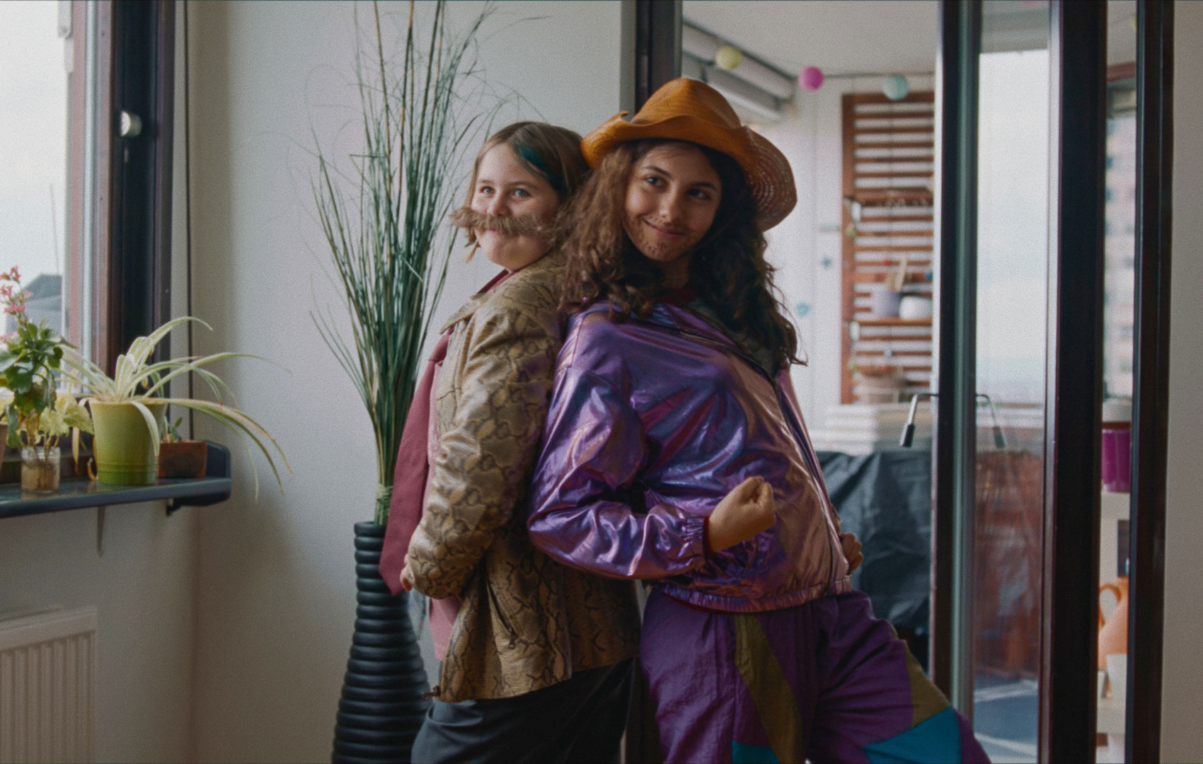 Zwei Teenagerinnen haben sich als Männer verkleidert und nehmen Rücken an Rücken stehend eine präsentierende Pose ein. Sie schauen selbstbewusst und glücklich.