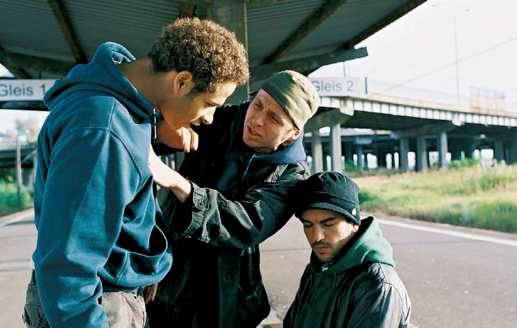 Drei männliche Jugendliche diskutieren hitzig miteinander unter einer Autobrücke. Einer sitzt, einer steht und redet auf den sitzenden ein. Der dritte scheint dazwischen zu gehen.